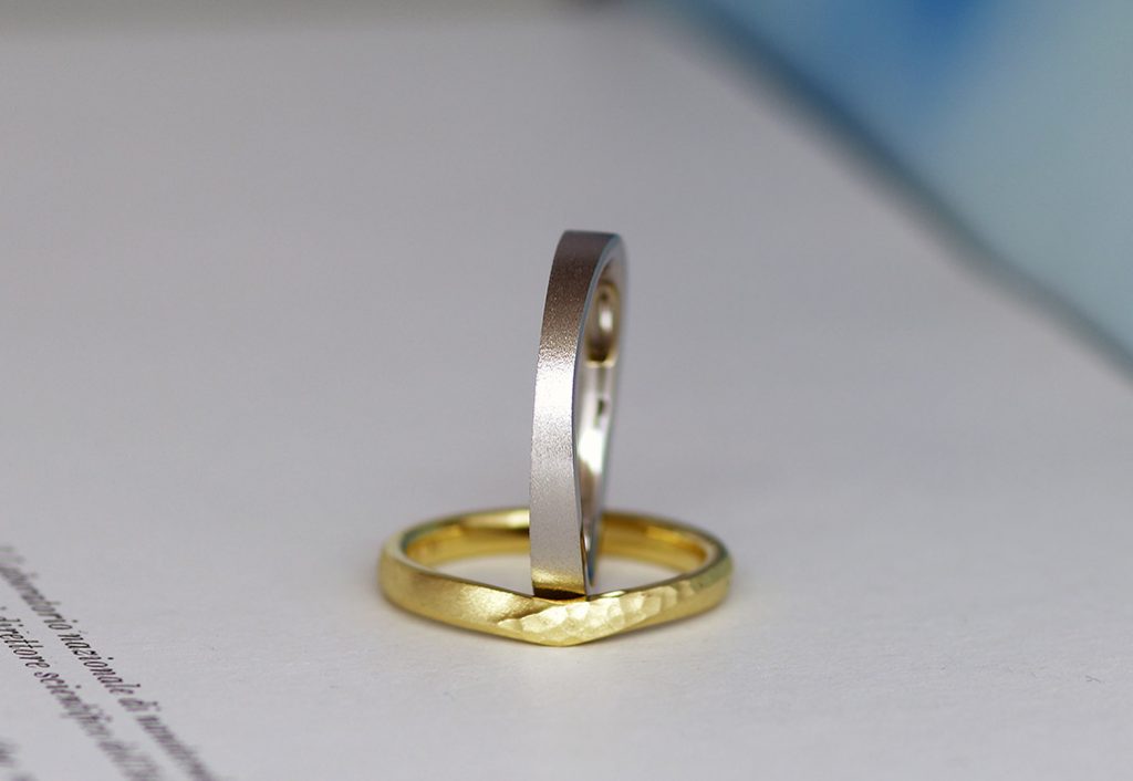 プラチナ素材でアシンメトリーデザインのメンズリングとV字にひねりと鎚目模様を入れたイエローゴールド素材のデザイン違いの結婚指輪（マリッジリング）